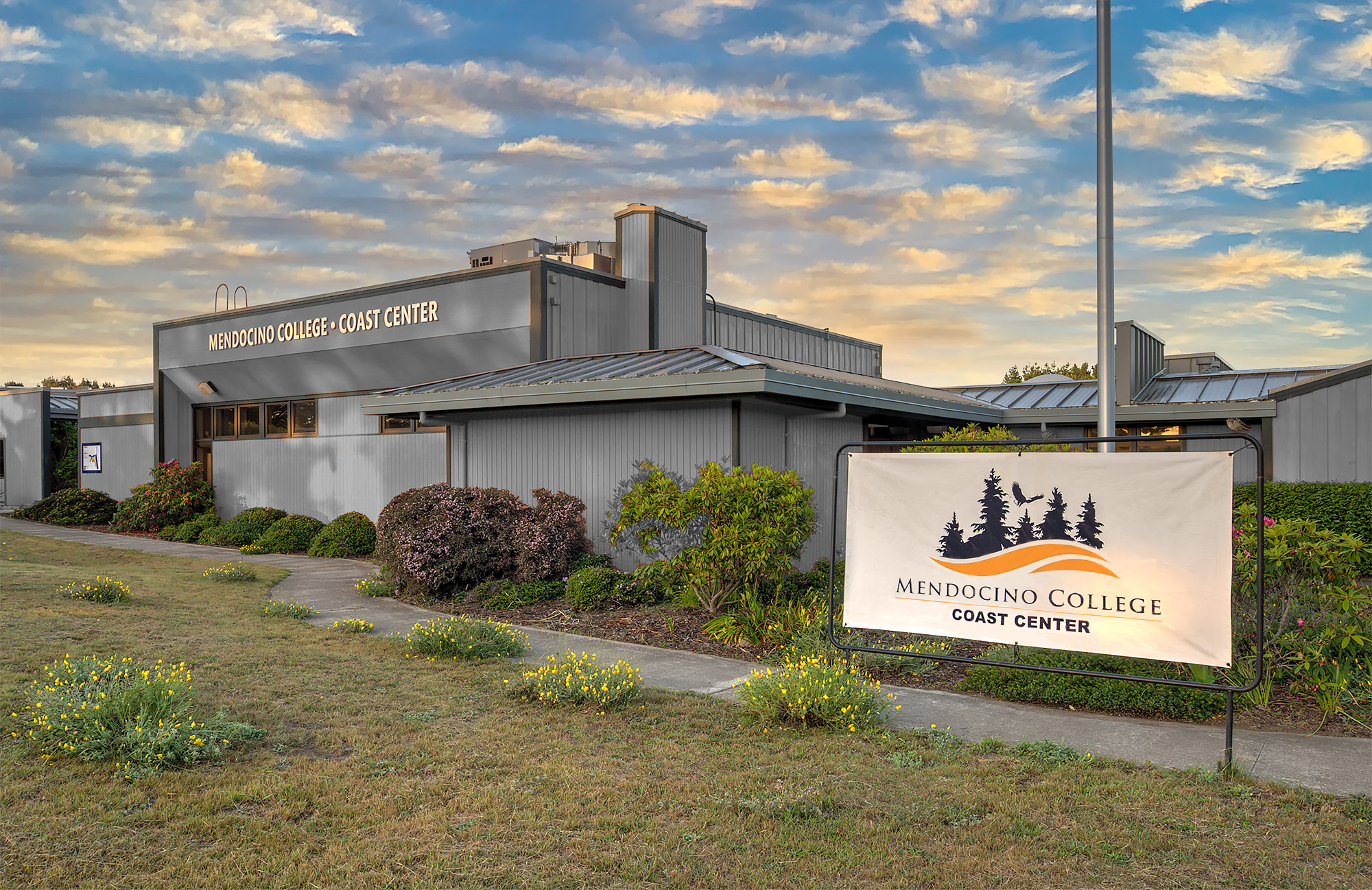 Photo of the front exterior of the Coast Center with signage and landscaping in front, setting sun and clouds in the background. 